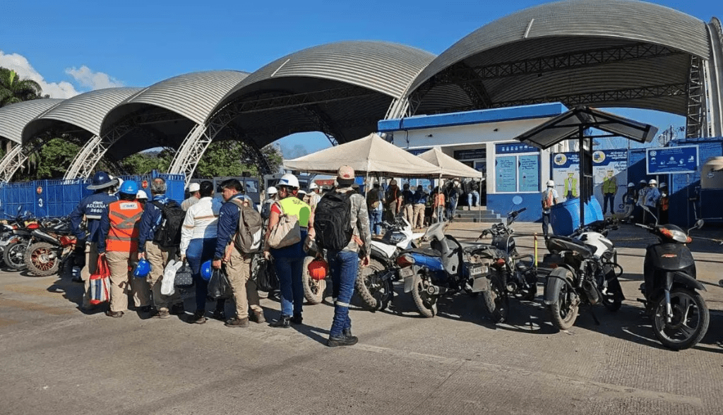 Personas en fila esperando ingresar a una instalación, con vehículos estacionados en primer plano y un cielo despejado al fondo.