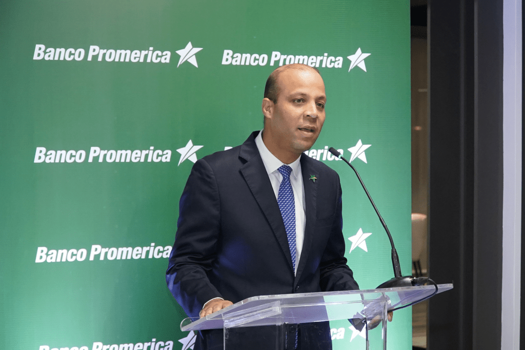 Un representante de Banco Promerica hablando en un evento con fondo verde con el logo del banco.