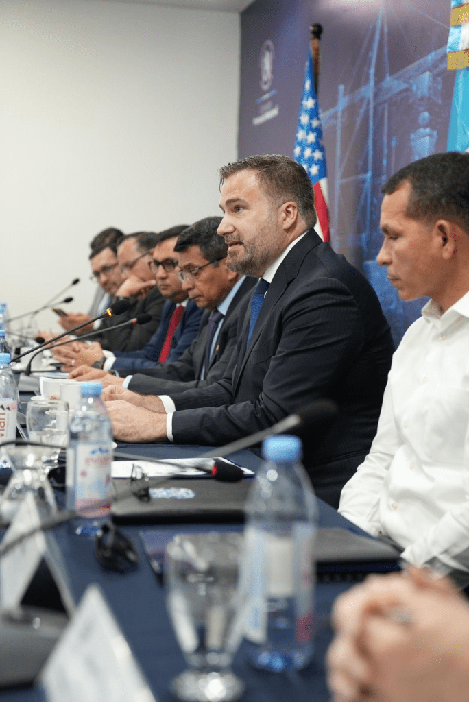 Reunión de la delegación de la Guardia Costera de Estados Unidos con autoridades guatemaltecas en una mesa de discusión, con banderas de Estados Unidos y Guatemala de fondo.