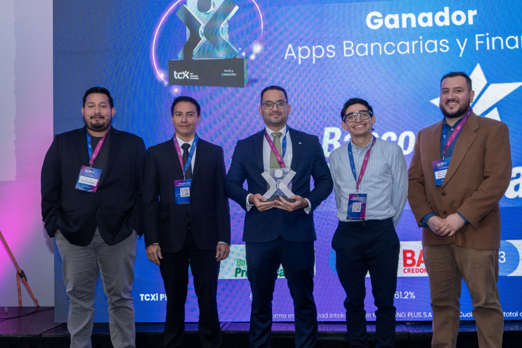 Grupo de cinco hombres de pie, celebrando la victoria del Banco Promerica en el certamen de Apps Bancarias y Financieras, sosteniendo un trofeo en el centro frente a un fondo decorado con luces y un logotipo del evento.
