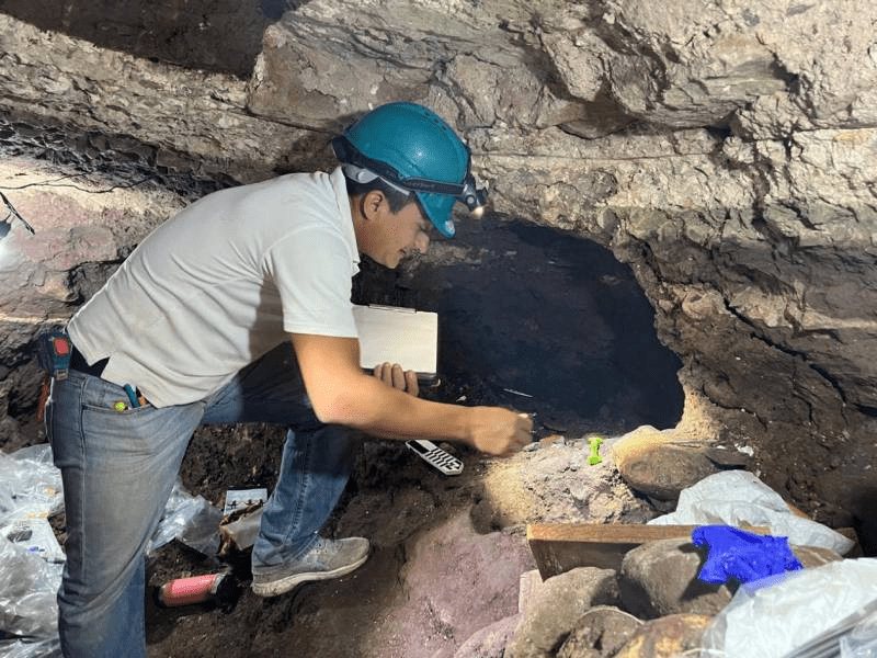 Un arqueólogo trabaja en un túnel del parque arqueológico de Copán, utilizando herramientas y tomando notas en un entorno de excavación.