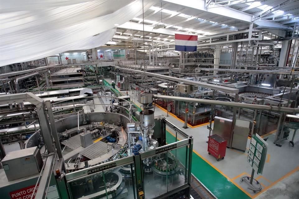 Vista interior de una planta de producción de cerveza, mostrando maquinarias y sistemas de transporte, con una bandera y equipos visibles en el entorno.