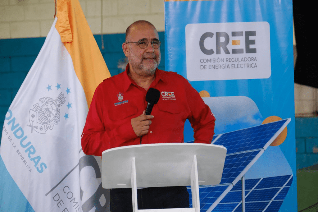 Funcionario de la Comisión Reguladora de Energía Eléctrica (CREE) de Honduras hablando en un evento, con bandera del país y un banner de la CREE al fondo.